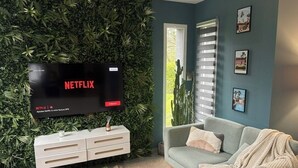 Un salon doté d’un téléviseur à écran plat diffusant Netflix, d’un meuble de rangement blanc, d’un canapé gris et d’un mur vert orné de plantes.