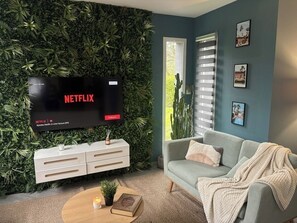 A living room with a flat-screen TV displaying Netflix, a white entertainment unit, a grey sofa, and a green wall with plants.