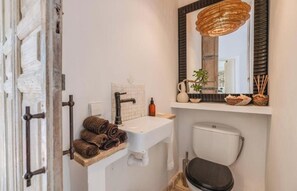 Ein Badezimmer mit weißem Waschbecken, Toilette, Spiegel mit Korbschirm und Ablagefläche mit Handtüchern und Pflanze.