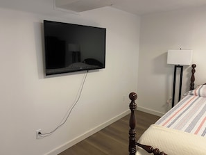 Ein Schlafzimmer mit an der Wand befestigtem Fernseher, einer Nachttischlampe, einem hölzernen Bettgestell und einer weißen Wand.