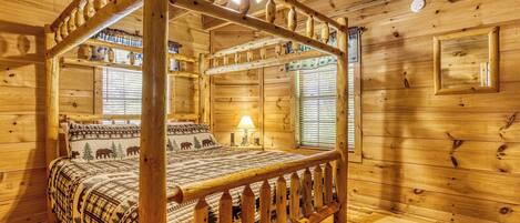 A wooden four-poster bed with a plaid bedspread, a wooden ceiling fan, and a window with blinds.