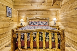 Ein Holzblockhaus-Innenraum mit einem Bett, das ein gestepptes Kopfstück, zwei Nachttischlampen und ein gerahmtes Bild an der Wand hat.