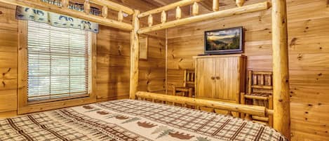 Habitación de cabaña de troncos con cama, ventana con persianas, ventilador de techo y un cuadro enmarcado en la pared.