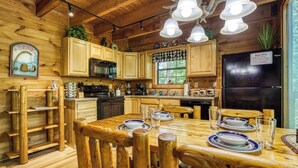 A wooden cabin interior with a dining table set for four, wooden cabinets, a black refrigerator, and a window with a view. - Mountain Views & Hot Tub | Near Dollywood (Sevierville)