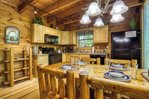 A wooden cabin interior with a dining table set for four, wooden cabinets, a black refrigerator, and a window with a view. - Mountain Views & Hot Tub | Near Dollywood (Sevierville)