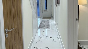 Un corridoio con una porta in legno, pavimento in marmo, illuminazione a incasso e una lampada.