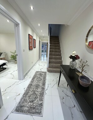 A hallway with a marble floor, a staircase, a black console table with decorative items, and framed pictures on the walls. - Spacious & Relaxing 4BR House | Excellent transpo. (Luton)