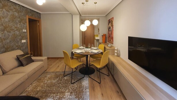 Een moderne woonkamer met een bank, eettafel, gele stoelen, een flatscreen televisie en een vloerkleed met een patroon.
