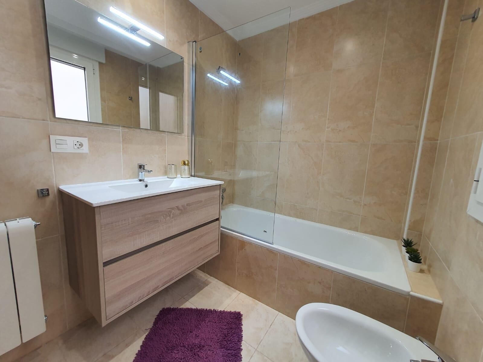 Een moderne badkamer met een wit wastafel, houten wastafelmeubel, een grote spiegel, een badkuip en een paars vloerkleed.