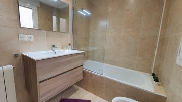 Een moderne badkamer met een wit wastafel, houten wastafelmeubel, een grote spiegel, een badkuip en een paars vloerkleed.