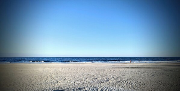 A beach with a clear sky, sandy shore, and a person walking.