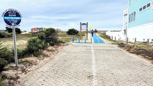 Un vialetto lastricato che conduce all'ingresso di una spiaggia, con un cartello di parcheggio, un edificio blu e una struttura in legno sullo sfondo.