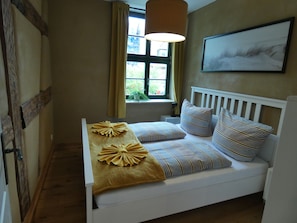 A bedroom with a white bed, striped pillows, a yellow throw, a window with curtains, and a painting on the wall.
