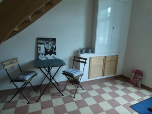 A room with a checkered floor, a table with chairs, a framed picture on the wall, a wardrobe, and a staircase.