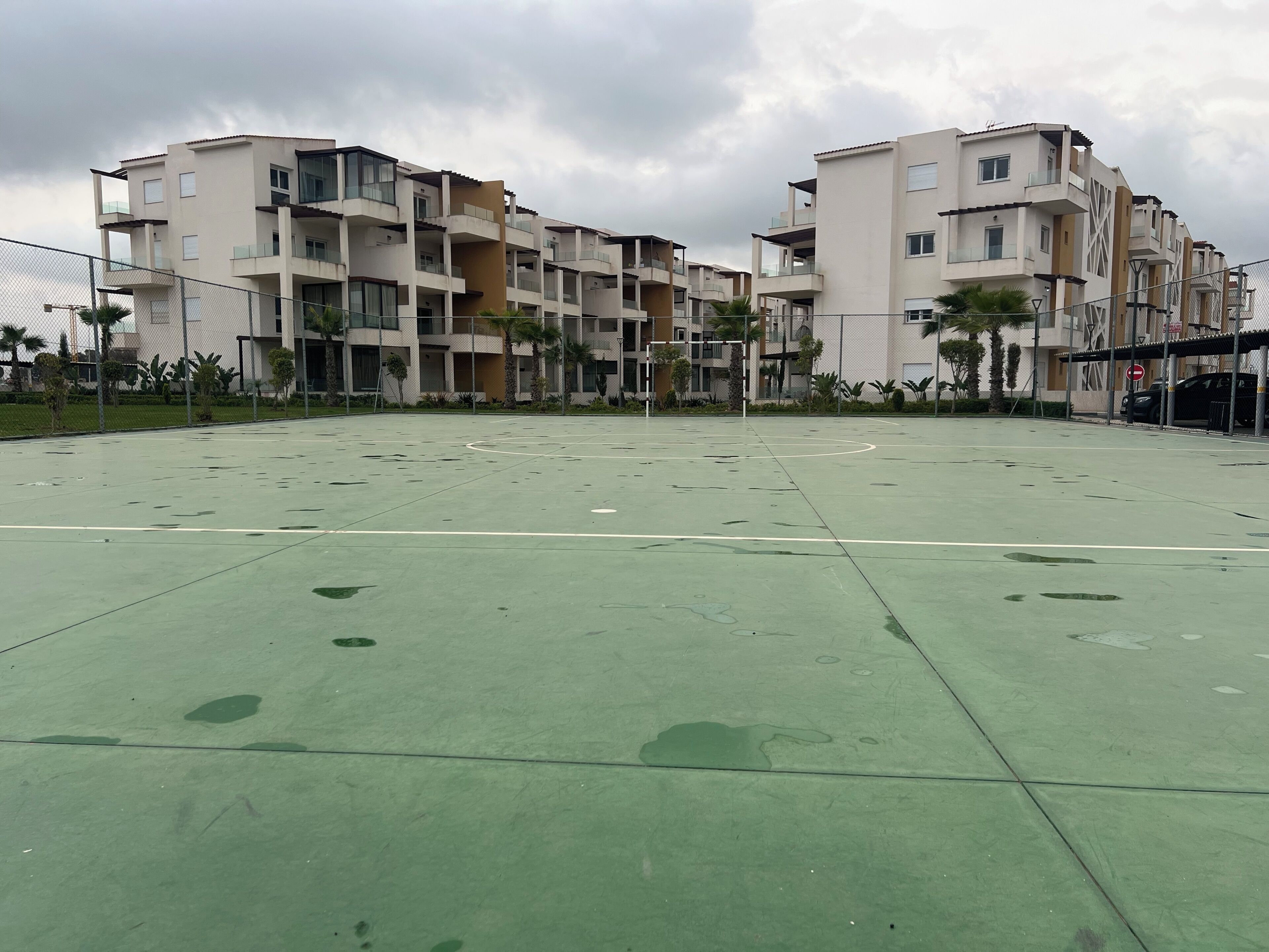 A multi-story residential building with balconies, a green sports court, and a clear sky.
