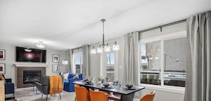 A modern living room with a fireplace, a dining area with orange chairs, a large window with curtains, and a mounted TV displaying 'NETFLIX'.