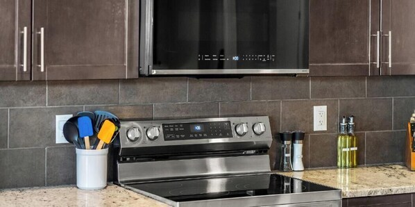 Una cocina con estufa de acero inoxidable, un microondas encima y un recipiente blanco para utensilios en la encimera.