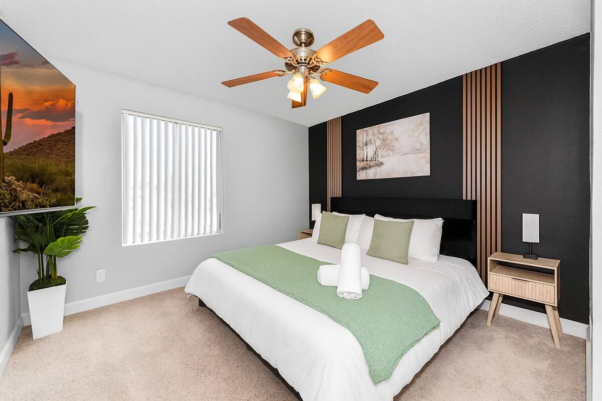 A bedroom with a bed, a ceiling fan, a window with blinds, a wall-mounted artwork, a bedside table, and a potted plant.