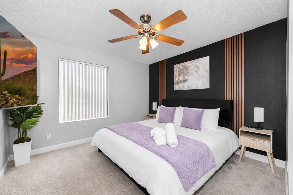 A bedroom with a bed, a ceiling fan, a nightstand, a wall-mounted lamp, and a window with blinds.