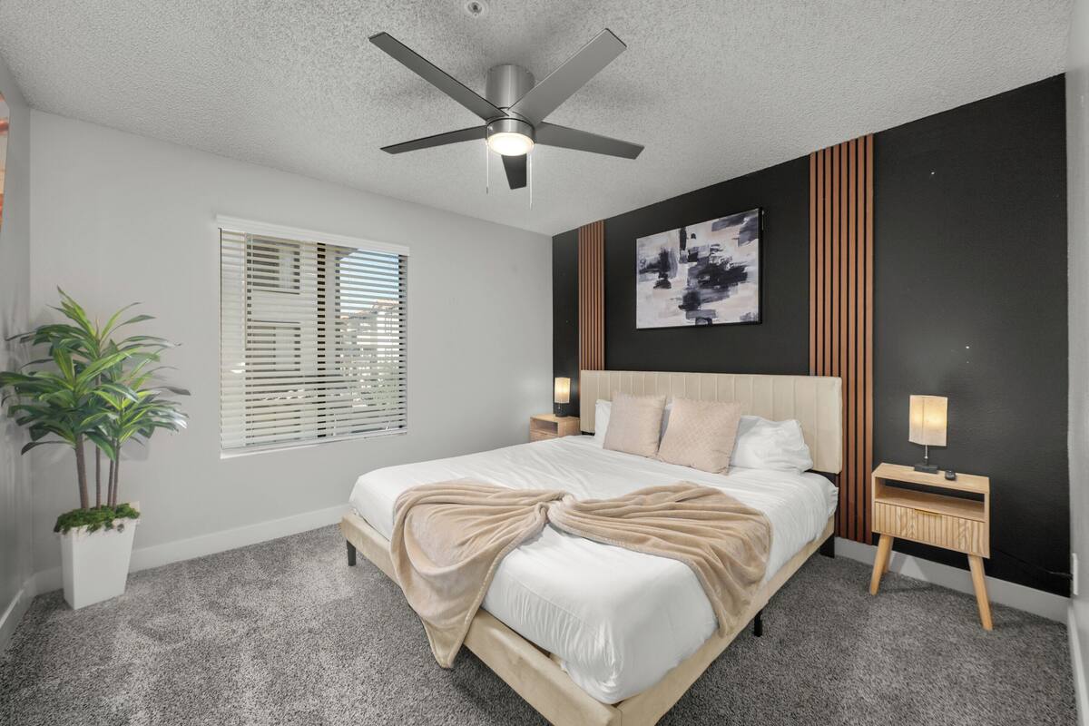 A bedroom with a bed, ceiling fan, bedside tables, a nightstand, a wall-mounted artwork, and a potted plant.