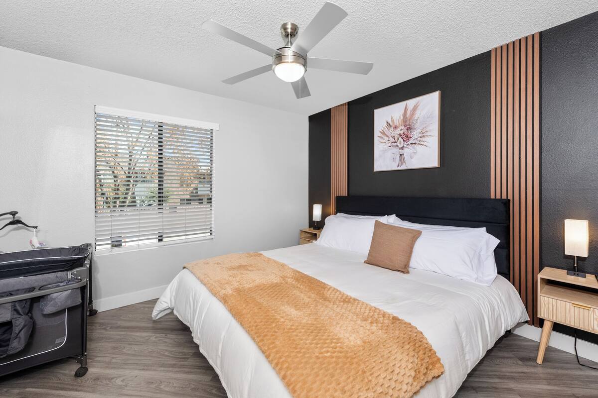 A bedroom with a bed, a ceiling fan, a nightstand, a lamp, a crib, and a window with blinds.