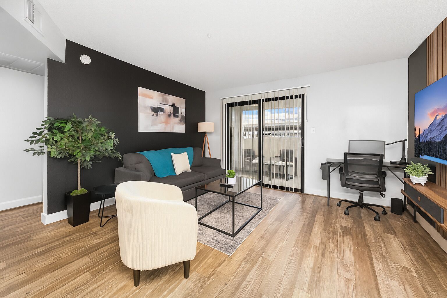 Un salon moderne comprenant un canapé, un fauteuil, une table basse, un bureau avec un ordinateur, une plante et une grande fenêtre.