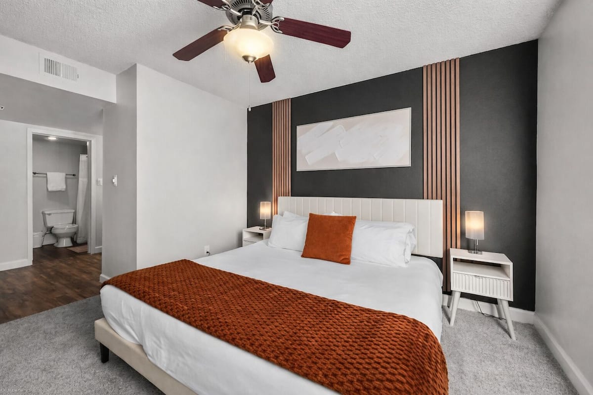 A bedroom with a bed, a ceiling fan, a nightstand, a wall-mounted lamp, and a bathroom visible in the background.