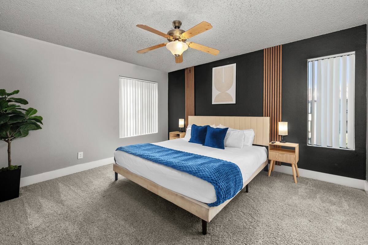 A bedroom with a bed, ceiling fan, bedside tables, lamp, and a potted plant.