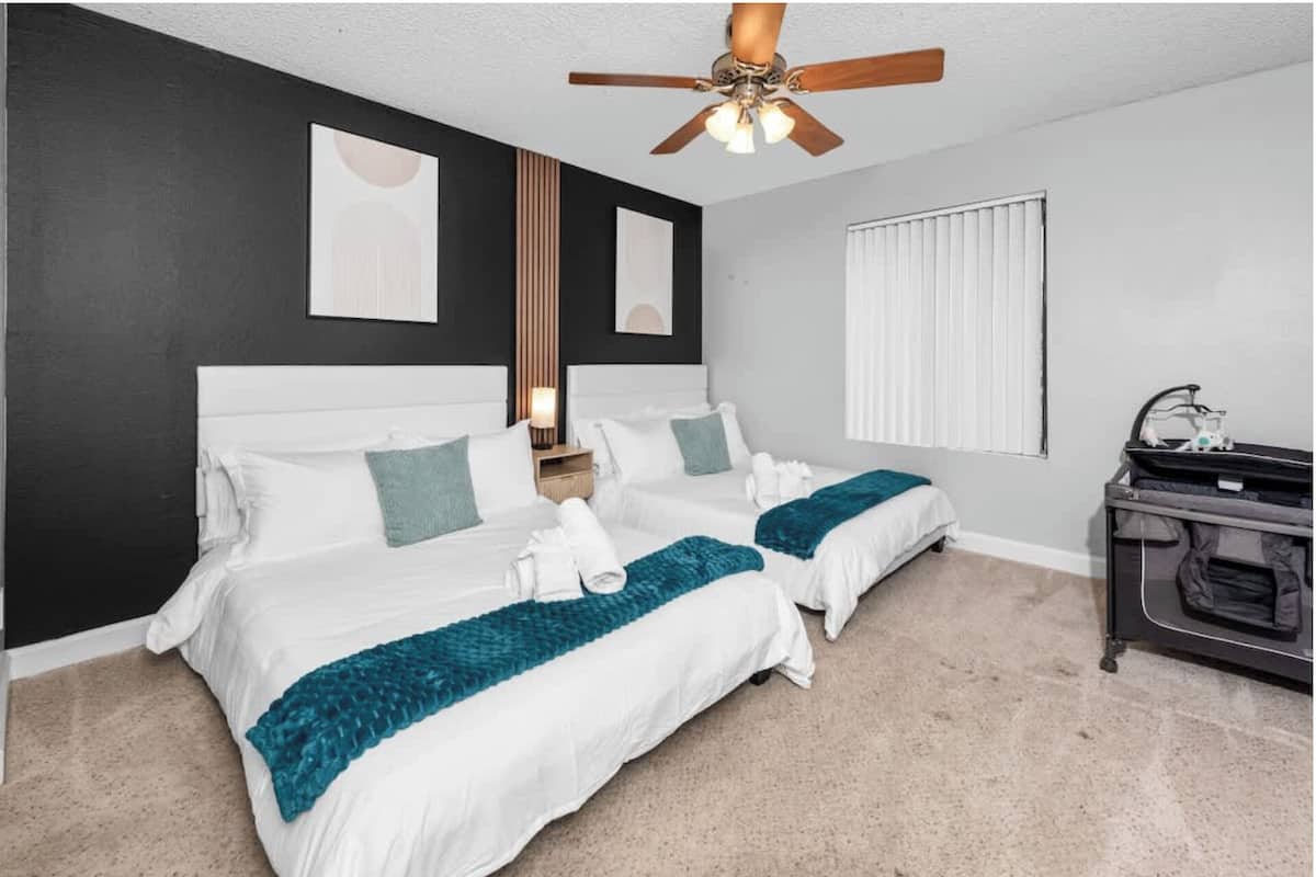 A hotel room with two beds, a ceiling fan, a window with blinds, and a small kitchenette.