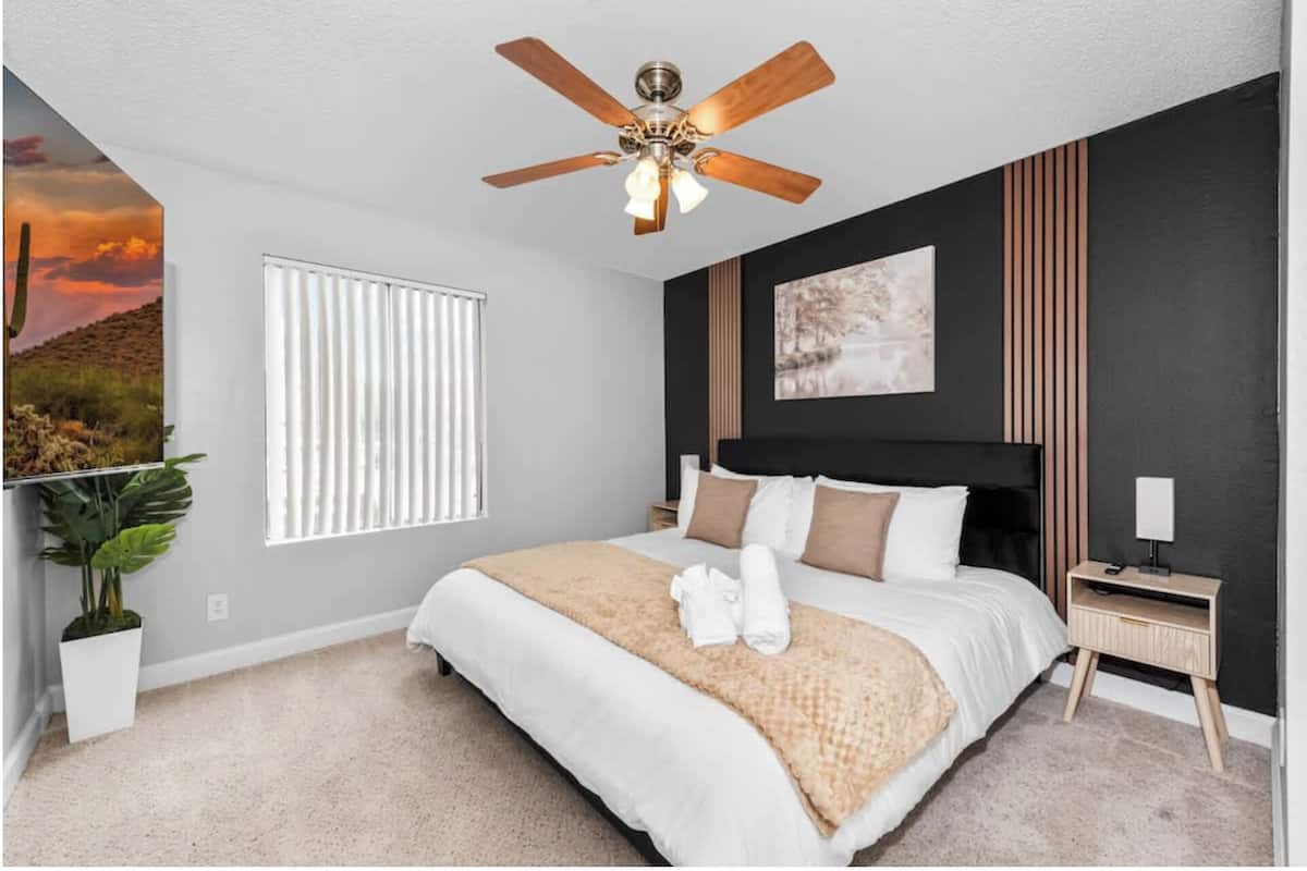 A bedroom with a bed, a ceiling fan, a window with blinds, a bedside table, and a wall-mounted artwork.