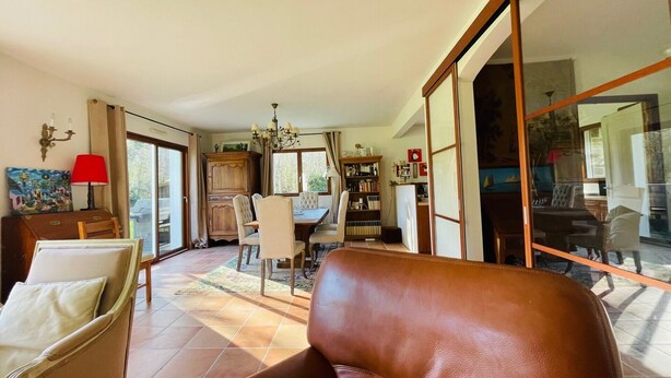 A spacious living room with a large brown leather sofa, a dining area with a table and chairs, a bookshelf, and a sliding glass door leading to a balcony. - Maison Familiale- Carnac & La Trinité (Carnac)