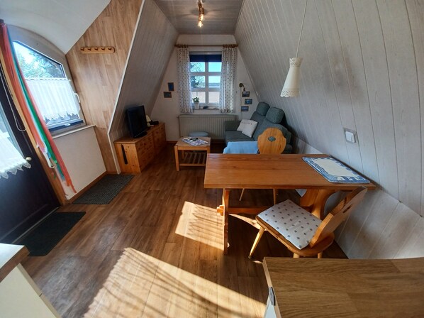 Ein gemütliches Dachzimmer mit Holztisch, einer Couch, einem Fernseher und einem Fenster mit Vorhängen.