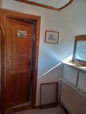 Une porte en bois avec une pancarte indiquant « WC », un tableau encadré accroché au mur et une fenêtre donnant sur un paysage.