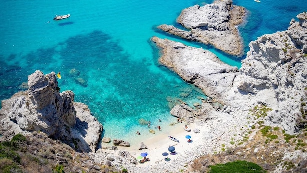 Un paysage côtier caractérisé par un terrain rocheux, des eaux turquoise et limpides, et une plage.
