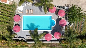 An aerial view of a swimming pool surrounded by lounge chairs and umbrellas, with greenery and a stone wall.