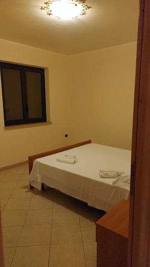A small, single-bed room with a window, a ceiling light, and a tiled floor.