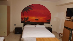 Une chambre à coucher comprenant un lit, des tables de chevet, une table en bois et une fresque murale représentant un coucher de soleil avec des palmiers et un navire.