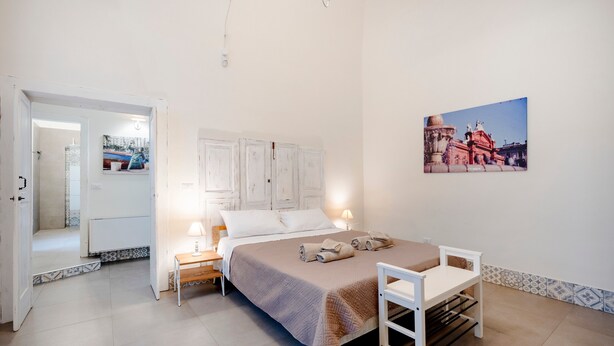 Ein Schlafzimmer mit einem Bett, Nachttischen, einem Sessel, einem Gemälde und einer Tür zu einem anderen Raum.
