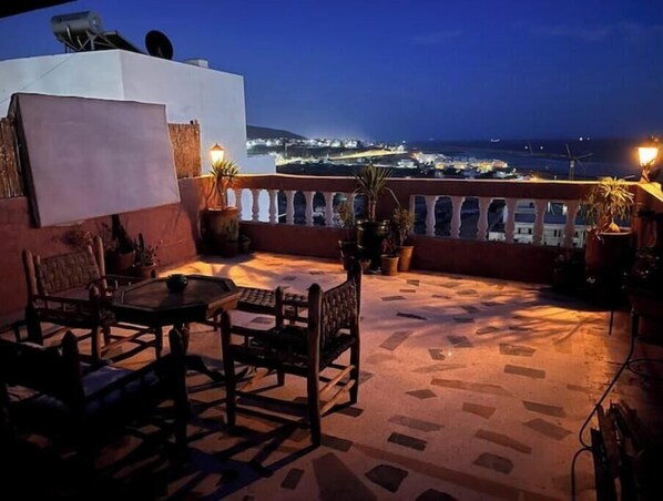 Eine Terrasse im Freien mit Tisch und Stühlen, Blumentöpfen und Blick auf eine Küstenstadt bei Nacht.
