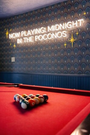 Ein Billardtisch mit Kugeln in der Mitte, eine Wand mit geometrischem Muster und ein Neon-Schild mit der Aufschrift 'NOW PLAYING: MIDNIGHT IN THE POCONOS'.