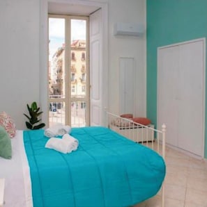 Un dormitorio con cama, ventana con vista a edificios, una puerta y una planta.