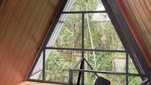 A room with a sloped ceiling, a large window with a view of greenery, and a bed with dark cushions. - Refugio de la Niebla Puyo Glamping (Puyo)
