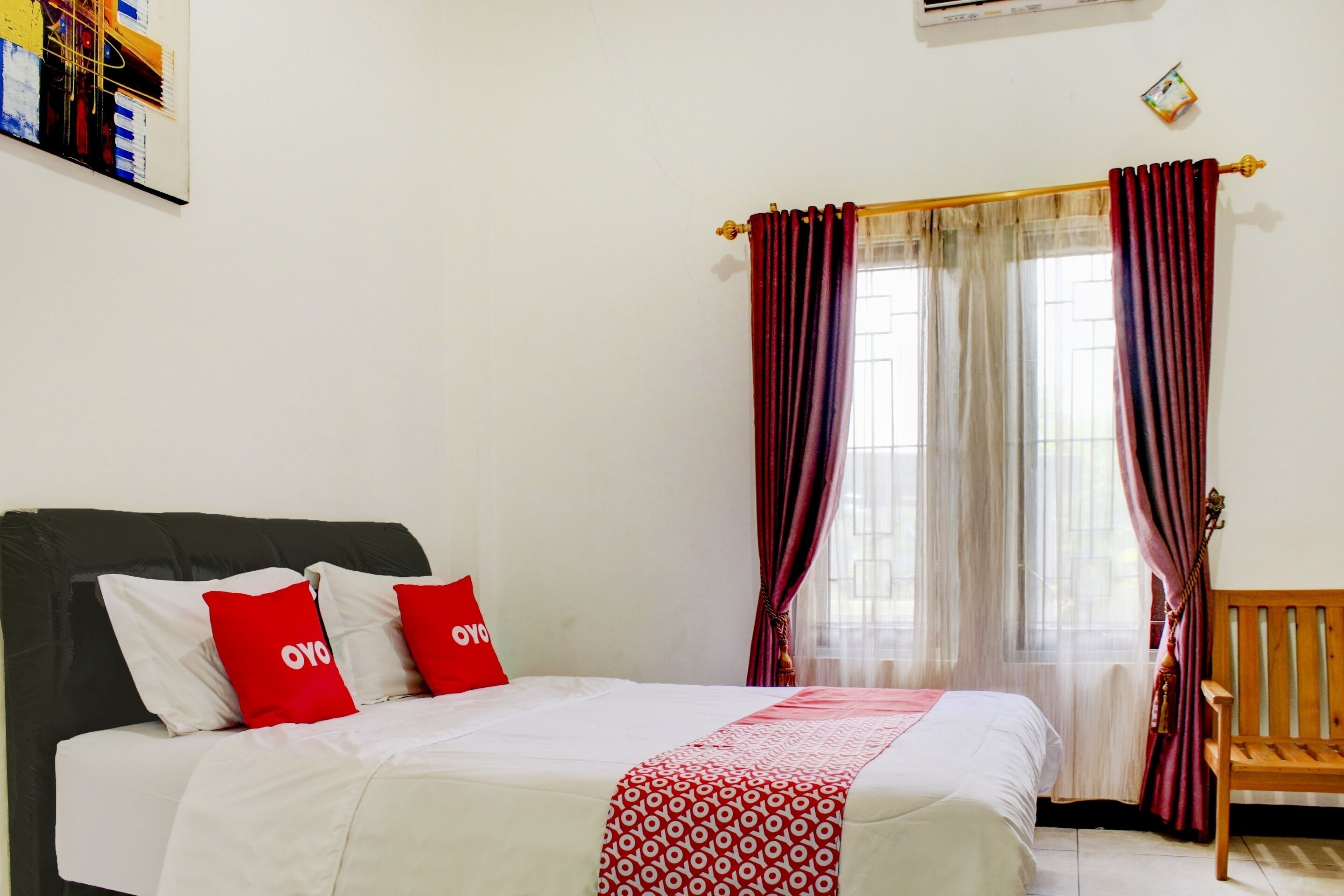 Un dormitorio con cama, una banca, una ventana con cortinas y un aire acondicionado instalado en la pared.