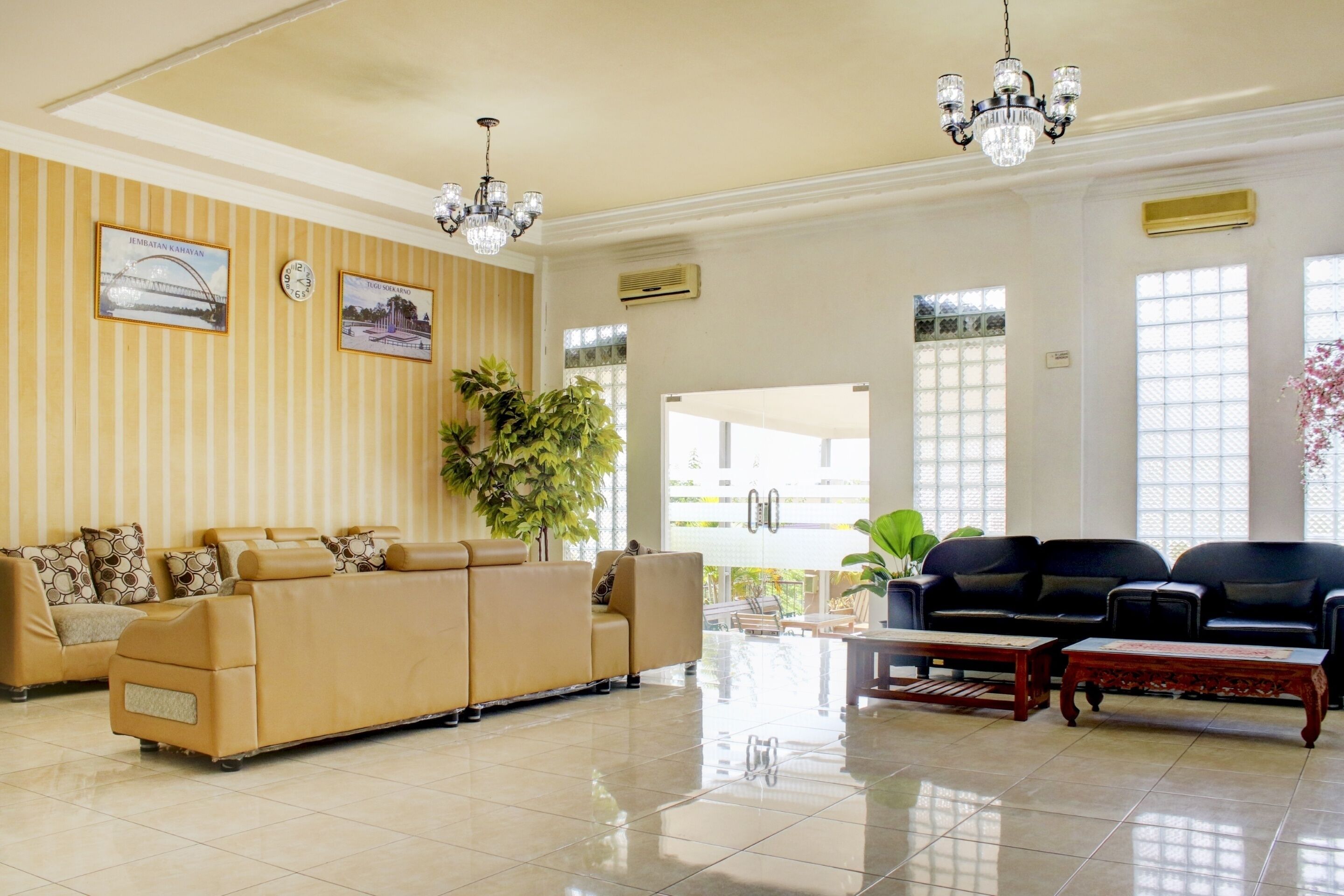 A spacious lobby with a beige sofa set, a black sofa set, a large potted plant, a chandelier, and a clock on the wall.
