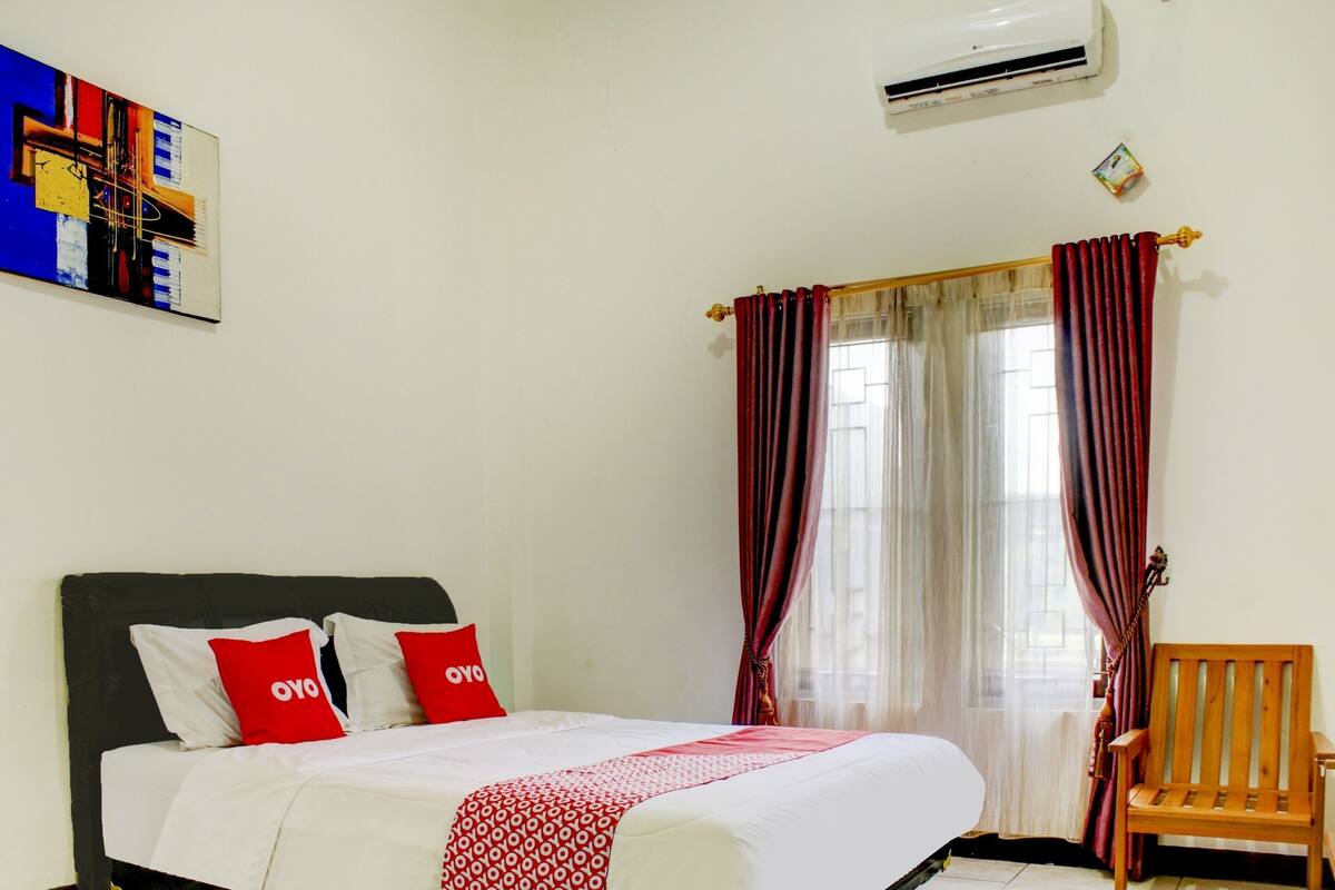 A hotel room with a bed, a chair, a painting, and a window with curtains.