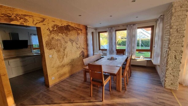 A dining area with a wooden table and chairs, a kitchen with a TV, and large windows with curtains. - Large vacation apartment with garden (Lauda-Königshofen)