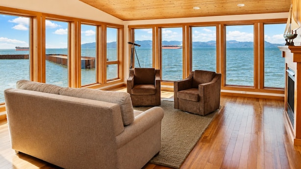 Un espacio amplio con techo de madera, ventanas grandes con vistas a un cuerpo de agua, un sofá, dos sillones y un telescopio.