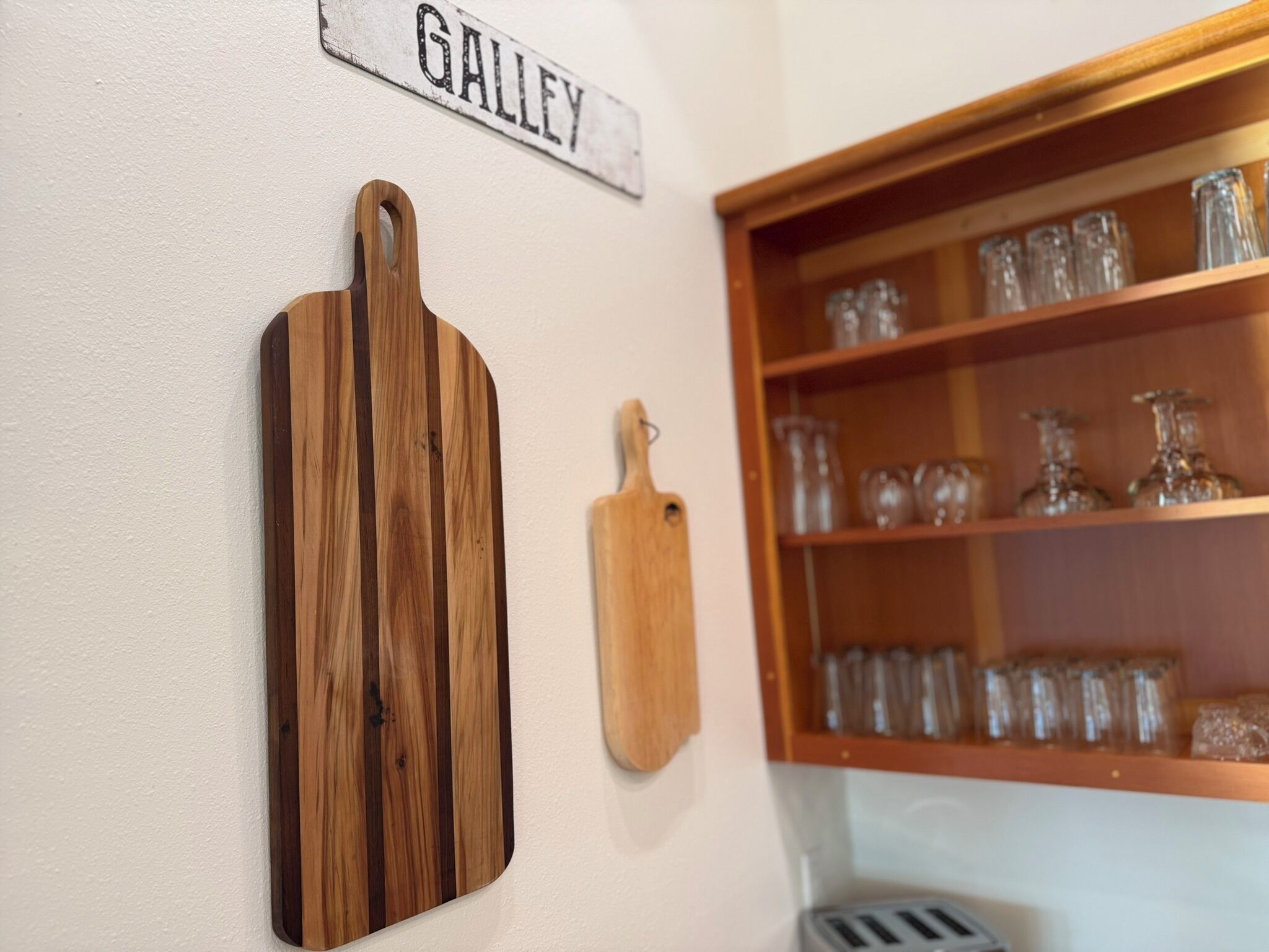 Una cocina con tabla de madera, cuchara de madera colgante y repisa con cristalería.