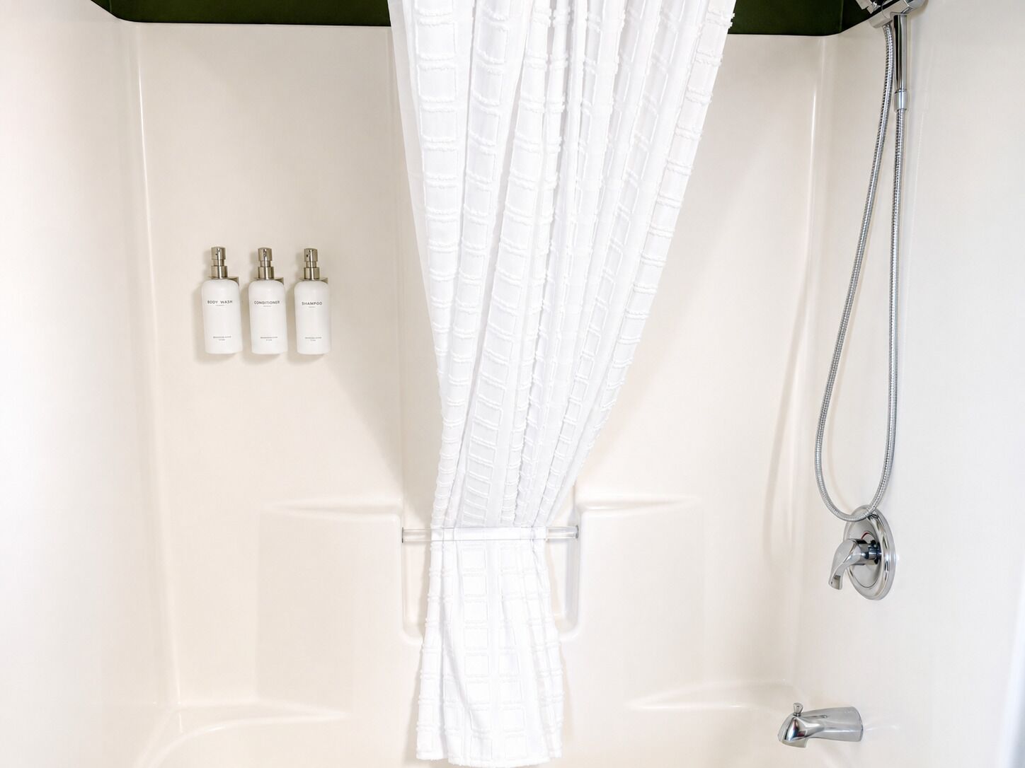 A white shower area with a shower curtain, a handheld showerhead, and three bottles of toiletries.