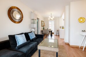 Ein modernes Wohnzimmer mit einem dunklen Sofa, einem Glastisch und einem Essbereich im Hintergrund.